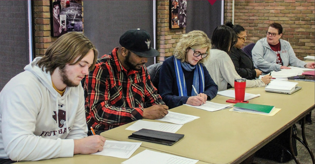 Students, faculty, and Maywood business owners working through materials together at a long table.